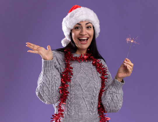 脖子快乐的年轻白人女孩戴着圣诞帽 脖子上戴着金箔花环 手里拿着节日火花灯 看着镜头 紫色背景上孤零零的空手帽子圣诞节年轻