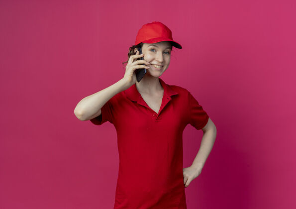 帽子身着红色制服 戴着帽子 面带微笑的年轻漂亮送货女孩在讲电话 手放在腰上 在深红色的背景下与复印空间隔离送货背景深红色