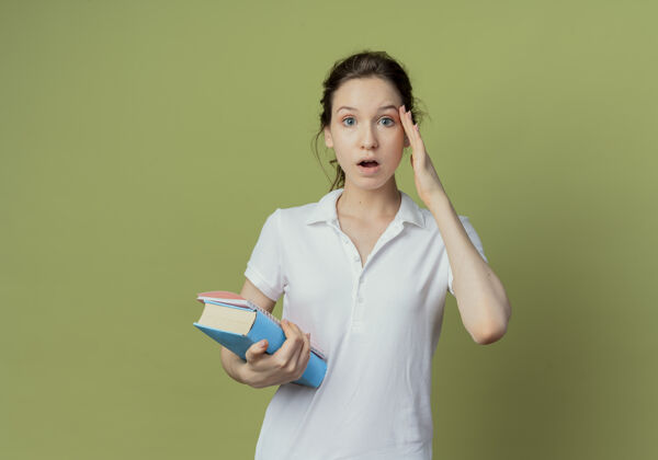 橄榄印象深刻的年轻漂亮的女学生拿着书和便笺簿把手放在脸上孤立的橄榄绿背景与复印空间印象手垫