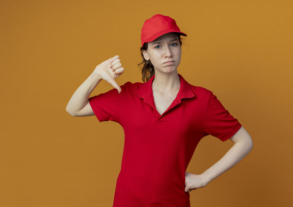 女孩年轻漂亮的送货员 穿着红色制服 戴着帽子 手放在腰上 拇指朝下 孤立地放在橙色背景上 留有复印空间红色年轻帽子