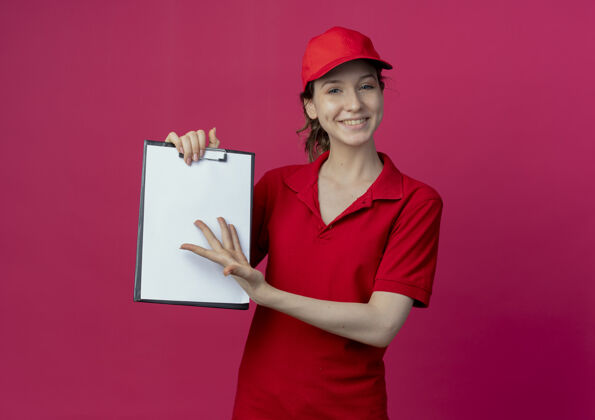 制服微笑着的年轻漂亮的送货女孩 穿着红色制服 戴着帽子 展示着剪贴板 用手指着它 在深红的背景上 有一个复制空间微笑交货年轻