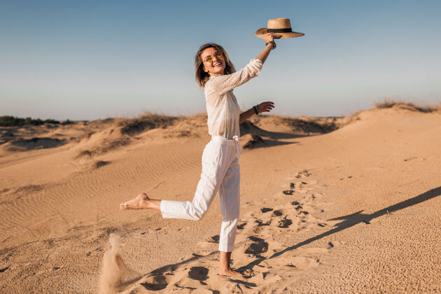跑步时髦的微笑美丽的快乐女人穿着白色的衣服在沙漠中奔跑和跳跃在日落时戴着草帽非洲裤子模特