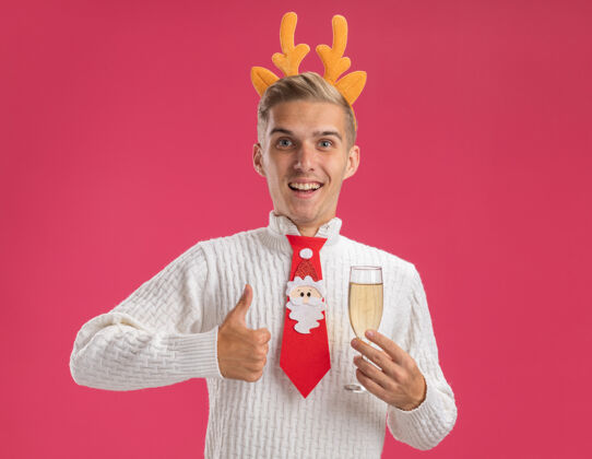 向上快乐的年轻帅哥戴着驯鹿鹿角头带 打着圣诞老人的领带 手里拿着一杯香槟 看着相机 在粉色背景上孤立地竖起大拇指粉红香槟年轻