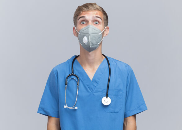 年轻惊讶地看着镜头年轻的男医生穿着医生制服听诊器和医用面罩隔离在白色的墙上站医生制服