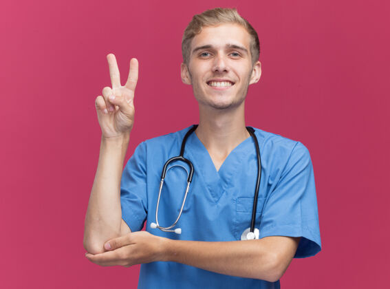 脸微笑的年轻男医生穿着医生制服 带听诊器 在粉红色的墙上显示出和平的姿态姿势表演衣服