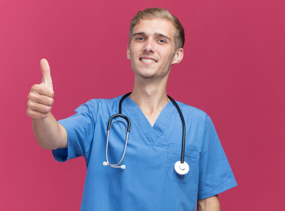 男性微笑着的年轻男医生穿着医生制服 听诊器在粉红色的墙上孤立地竖起大拇指感觉拇指公民