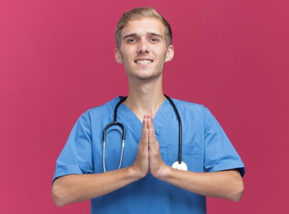 人面带微笑的年轻男医生穿着医生制服 听诊器显示祈祷手势 隔离在粉红色的墙上感觉人姿势