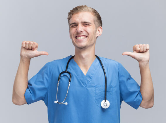 站着面带微笑的年轻男医生穿着医生制服 手持听诊器指着隔离在白墙上的自己微笑点人
