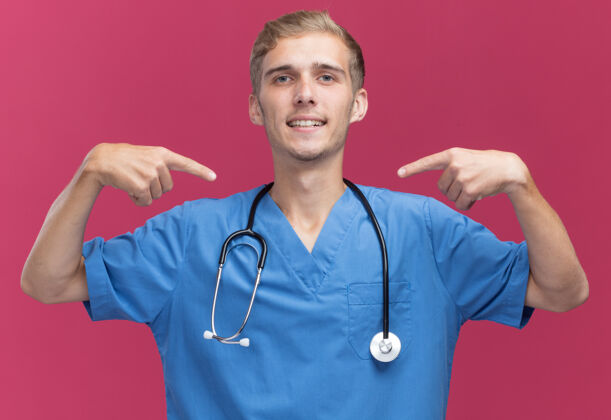 脸面带微笑的年轻男医生穿着医生制服 手持听诊器指着隔离在粉红色墙上的自己站着姿势听诊器
