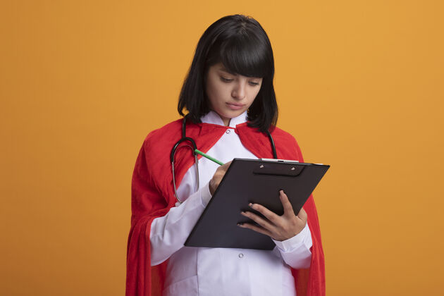 女人自信的年轻超级英雄女孩戴着听诊器 穿着医用长袍和斗篷 在剪贴板上写着什么穿长袍医生