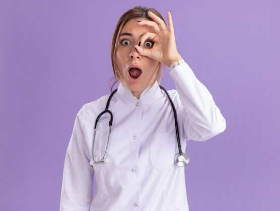 人惊讶的年轻女医生穿着医用长袍 听诊器显示出隔离在紫色墙上的表情衣服人医学