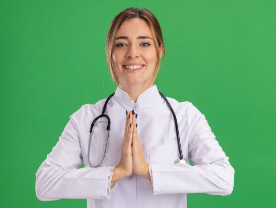 站着微笑的年轻女医生穿着医用长袍 听诊器在绿色的墙上显示着祈祷的手势女性祈祷表情