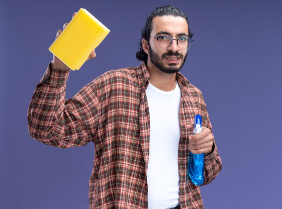 年轻自信的年轻帅气的清洁工穿着t恤拿着喷雾瓶用海绵隔离在蓝色的墙上瓶子清洁海绵