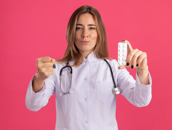 年轻年轻严格的女医生 穿着医用长袍 手持听诊器 手持药片 手持拳头 隔离在粉红色的墙上药丸严格女性