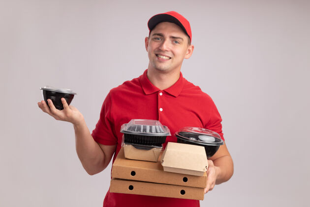 容器面带微笑的年轻送货员穿着制服 戴着帽子 把食物容器放在隔离在白墙上的比萨饼盒上披萨食物拿着