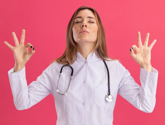 长袍年轻的女医生 闭着眼睛 穿着医用长袍 手持听诊器 显示冥想姿势 隔离在粉红色的墙上冥想显示女性