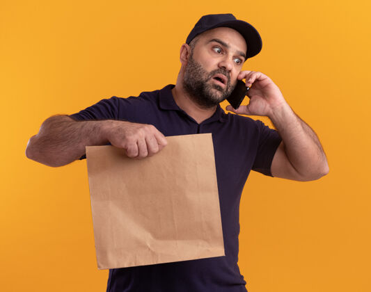 拿着惊恐地看着身边穿着制服 戴着帽子的中年送货员拿着纸食品包讲着电话隔离在黄色的墙上包装男人说话