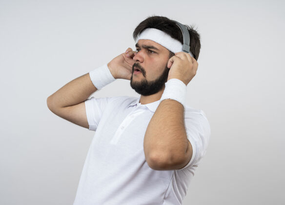 头带年轻的运动型男子一边看着戴着头带和腕带的耳机 一边隔离在白墙上 留着复印空间腕带耳机男士