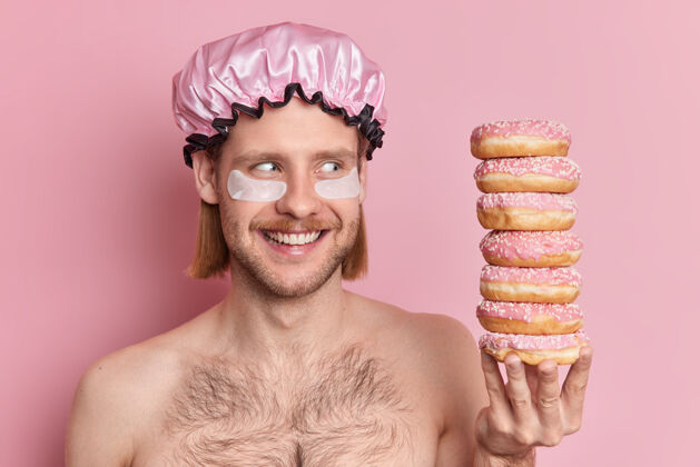 卫生室内拍摄的高兴的欧洲年轻人笑容满面看着一堆美味的甜甜圈有诱惑吃甜点摆出赤裸上身的姿势堆帽子赤膊上阵