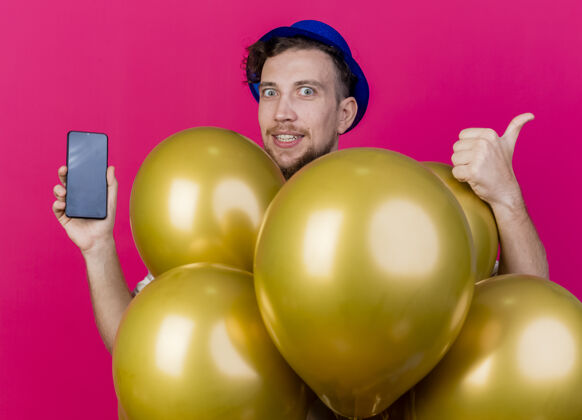 气球令人印象深刻的年轻英俊的斯拉夫党的家伙戴着党的帽子站在气球后面看前面显示手机和拇指向上孤立的粉红色墙上表演拇指脸