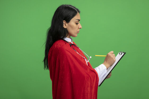 超级英雄站在侧视图年轻的超级英雄女孩穿着医用长袍 手持听诊器 在绿色的剪贴板上写着什么年轻穿立场