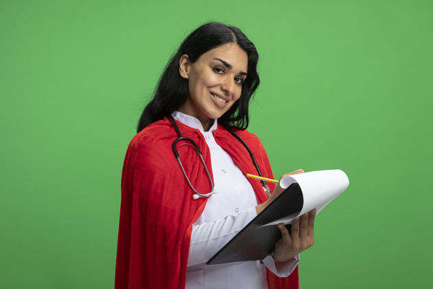 绿色微笑着的年轻超级英雄女孩 穿着医用长袍 手持听诊器 笔直地看着前方 在绿色的剪贴板上写着什么年轻什么写作
