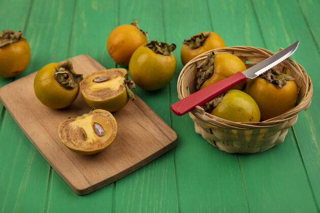 桶柿子水果的顶视图在一个桶上 在一张绿色的木桌上 用刀把柿子水果切成两半放在一个木制的厨房板上绿色木头刀