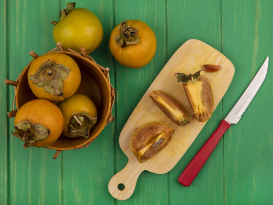 食物木制厨房板上的柿子水果减半俯视图 绿色木桌上的木桶上有一把柿子水果刀视野柿子一半