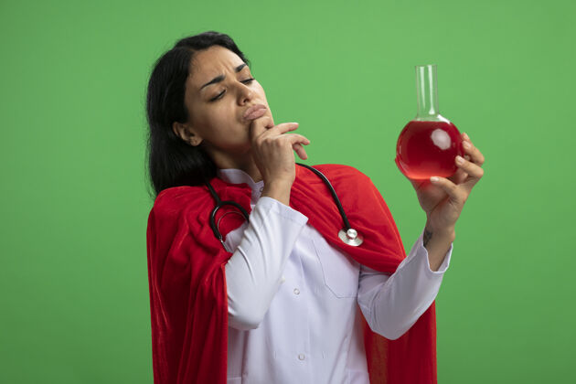 穿沉思的年轻超级英雄女孩穿着医用长袍 手持听诊器 看着装满红色液体的化学玻璃瓶 抓住绿色的下巴红色化学超级英雄