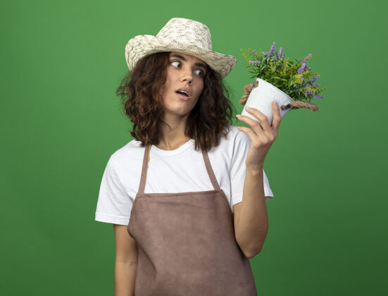女性惊讶的年轻女园丁穿着制服 戴着园艺帽 手持花盆 看着隔离在绿色花坛上的花朵惊喜花穿