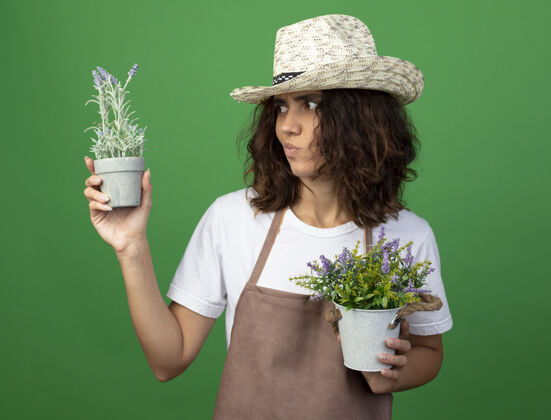 绿色沉思的年轻女园丁 穿着制服 戴着园艺帽 手拿花盆 看着隔离在绿色上的花朵花盆女性思想