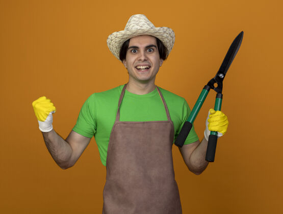 花园年轻的男园丁面带微笑 身着制服 戴着园艺帽 戴着手套 拿着剪刀 在橘色上显示“是”的手势穿剪子制服
