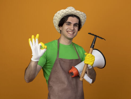 扩音器身着制服的年轻男园丁面带微笑 戴着园艺帽 戴着手套 拿着带耙子的扩音器 在橙色上显示“是”的手势穿园艺是的