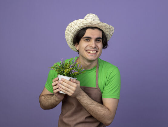 戴着微笑着的年轻男园丁穿着制服 戴着园艺帽 手里拿着花盆里的花制服微笑人
