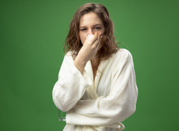 疾病虚弱的生病的年轻女孩直直地看着前方 穿着白色长袍 用隔离在绿色上的餐巾擦鼻子虚弱鼻子穿