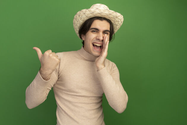 帅哥微笑着的年轻帅哥戴着帽子 在绿色的墙上喊着某人竖起大拇指拇指秀帽子