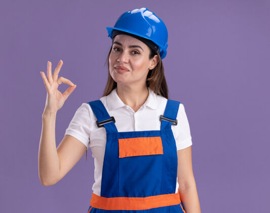 制服一个穿着制服的年轻建筑女在紫色的墙上展示了一个很好的姿势建筑工人手势女人