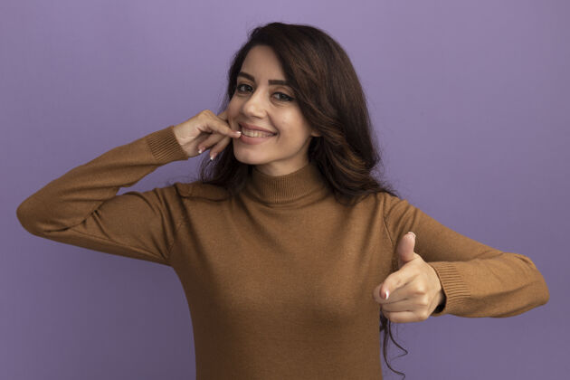 穿微笑着的年轻漂亮女孩穿着棕色高领毛衣 在紫色的墙上显示电话的手势和孤立点电话电话点
