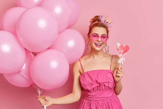 甜点积极的红发女孩在流行的粉红色阴影和礼服举行美味的糖果和一堆气球有节日气氛的聚会姿势在玫瑰色的背景下美味乐观一个