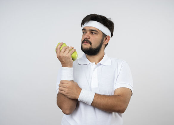 年轻自信的年轻运动型男子一边看着戴着头带和腕带的苹果 一边抓住手臂 隔离在白色上 留有复制空间运动手臂信心