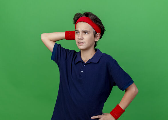 感情困惑的年轻英俊的运动男孩戴着头带和护腕 戴着牙套 手放在腰上 头后面 在绿色的墙壁上看着笔直的孤立的复制空间穿帅气腰