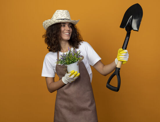 手套身着制服的年轻女园丁面带微笑 戴着园艺帽和手套 手里拿着花盆里的花 看着手中的铲子园艺女人手