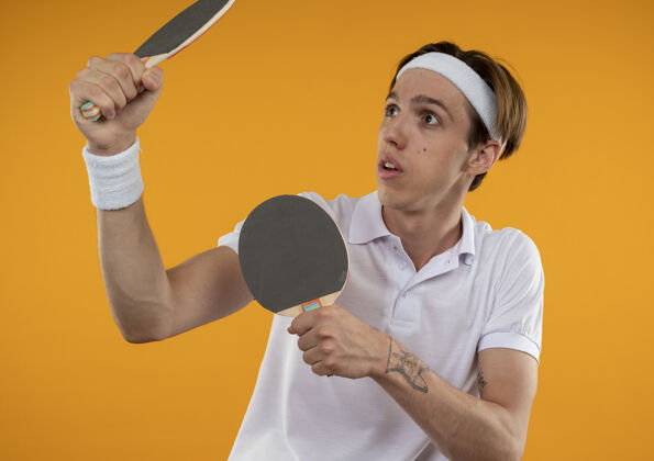 乒乓球惊讶的年轻人看着一边戴着头带和腕带举起乒乓球拍隔离在橙色的墙上穿小伙子壁板