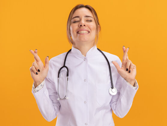听诊器微笑着闭上眼睛年轻的女医生穿着医用长袍听诊器交叉手指隔离在黄色的墙上微笑十字架医疗