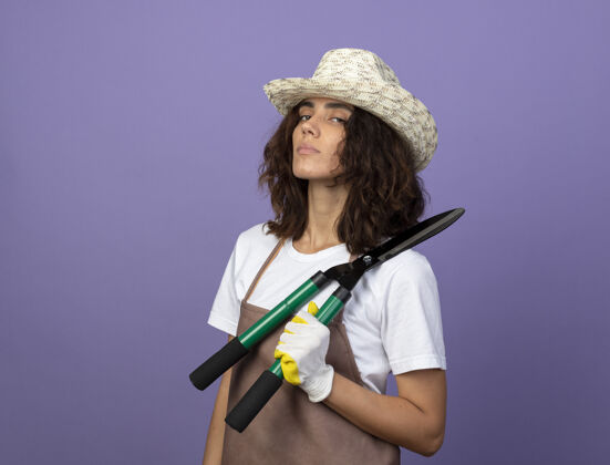 人自信的年轻女园丁穿着制服 戴着园艺帽和手套 肩上拿着剪刀紫色女士花园