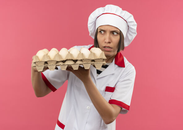 鸡蛋穿着厨师制服的年轻女厨师拿着一盒鸡蛋 看着粉红色背景上的鸡蛋不愉快烹饪纸箱