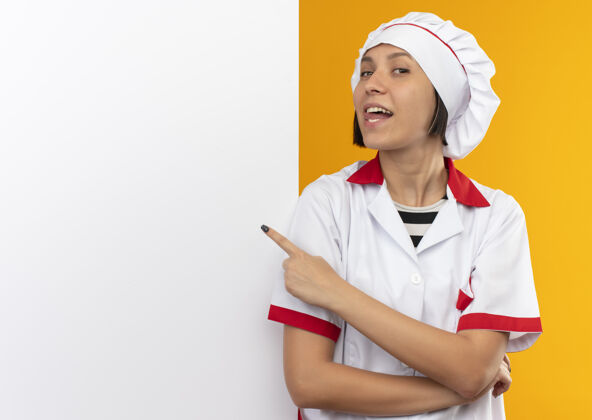 制服身着厨师制服的快乐年轻女厨师站在白色墙壁前 露出舌头 指着隔离在橙色墙上的墙壁欢乐站立厨师