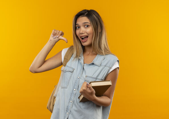 拿着印象深刻的年轻漂亮的学生女孩穿着背袋拿着打开的书指着自己孤立在橙色印象学生橙色