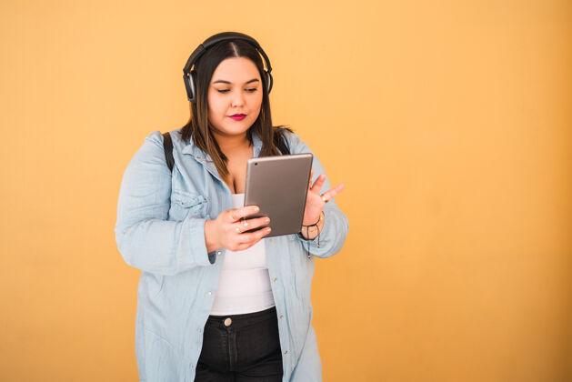 设备照片中的年轻女子戴着耳机和数字平板电脑在户外靠着黄色的墙壁听音乐耳机无线曲线