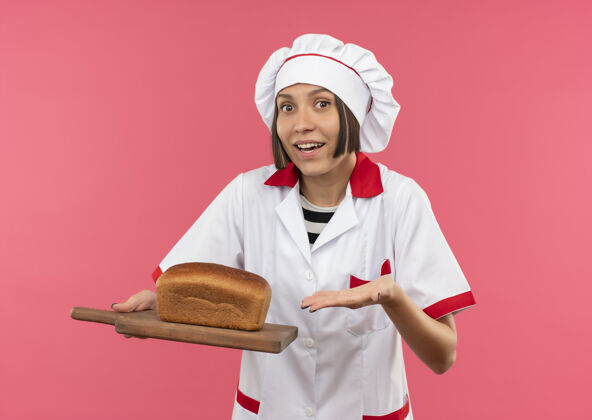 厨师身着厨师制服的快乐年轻女厨师手拿并指着插着粉红色面包的砧板董事会削减面包